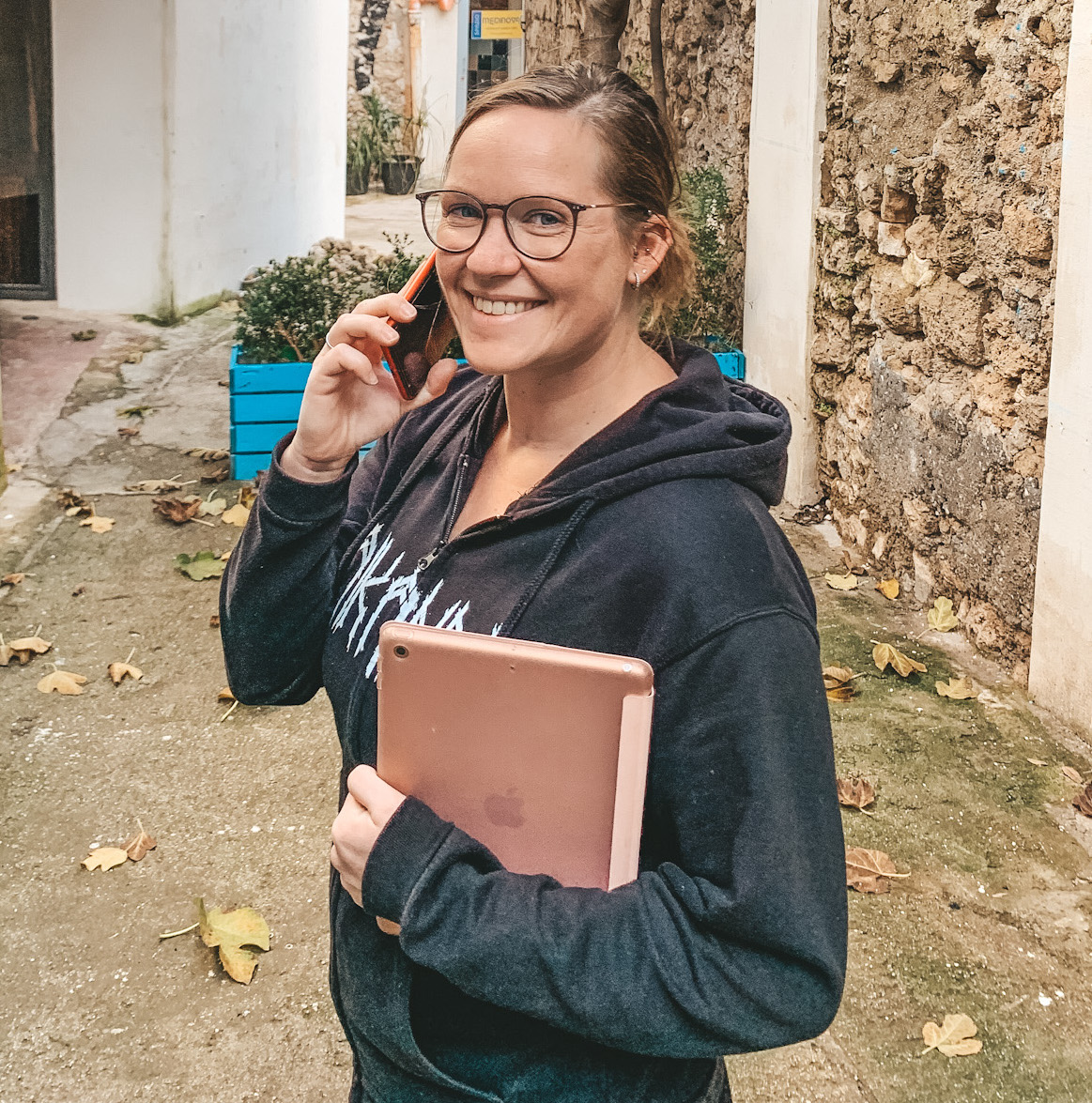 Kelly Prato mit Handy am Ohr und Tablet in der Hand vor einem schönen, rustikalen Hintergrund